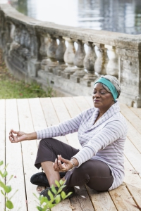 Portrait of a senior African American woman meditating, in yoga lotus pose. She is sitting on a deck by the water, legs crossed, with her eyes closed and a peaceful expression on her face.